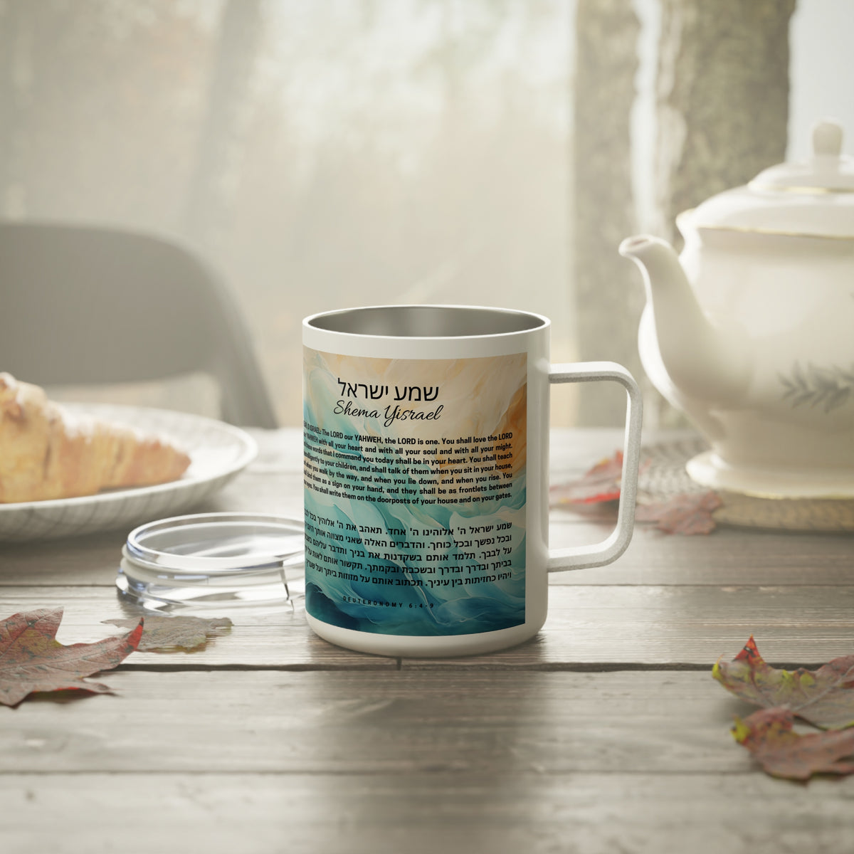 A white stainless steel coffee mug with a custom printed design featuring Hebrew text and a scenic landscape, shown with its lid on a rustic wooden table next to a white teapot.