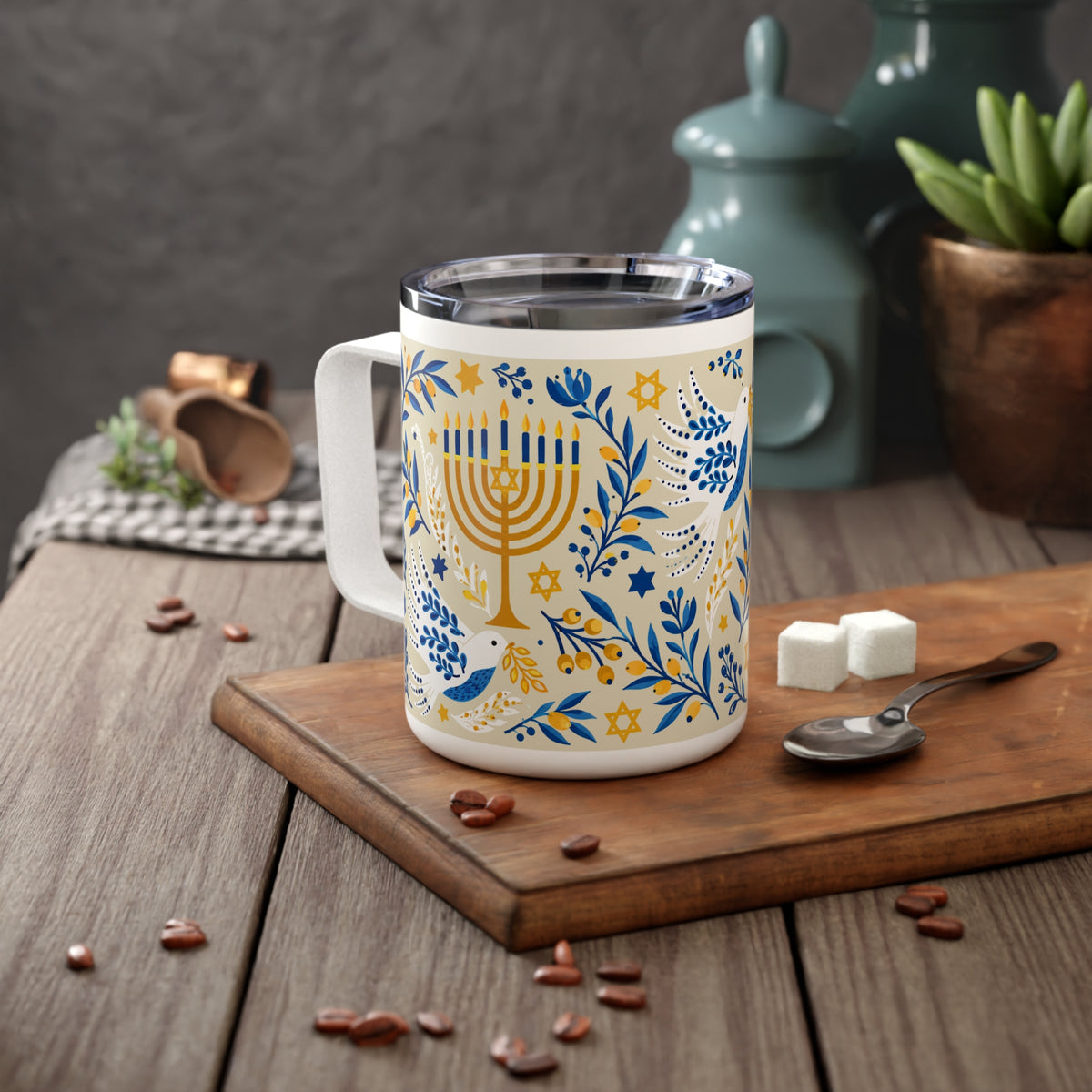 A white stainless steel mug with a printed blue and gold Hanukkah menorah design, set on a wooden cutting board with coffee beans, sugar cubes, and a spoon on a rustic wooden table.