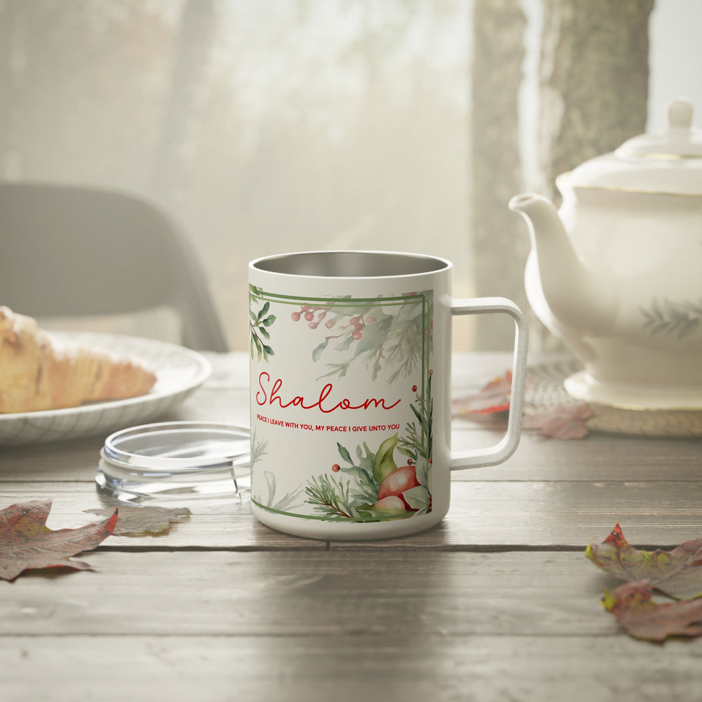 A white stainless steel  mug with a silver interior, featuring a green botanical border and the word 'Shalom' in red script, set on a rustic wooden table with a glass lid, a teapot, and autumn leaves in the background.
