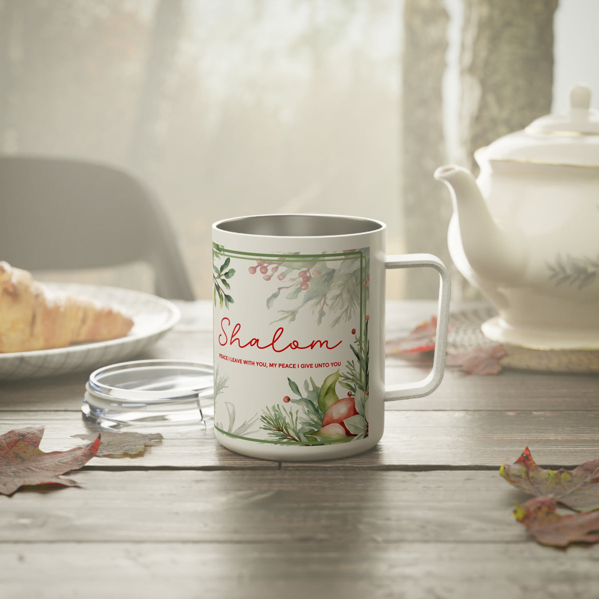 A white stainless steel  mug with a silver interior, featuring a green botanical border and the word 'Shalom' in red script, set on a rustic wooden table with a glass lid, a teapot, and autumn leaves in the background.