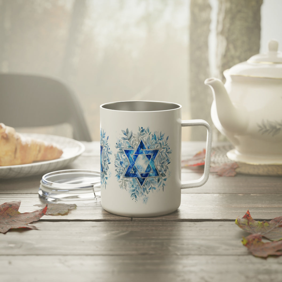 A white stainless steel mug with a blue Star of David and floral snowflake design, set on a rustic wooden table with a glass lid, a teapot, and a pastry in the background.