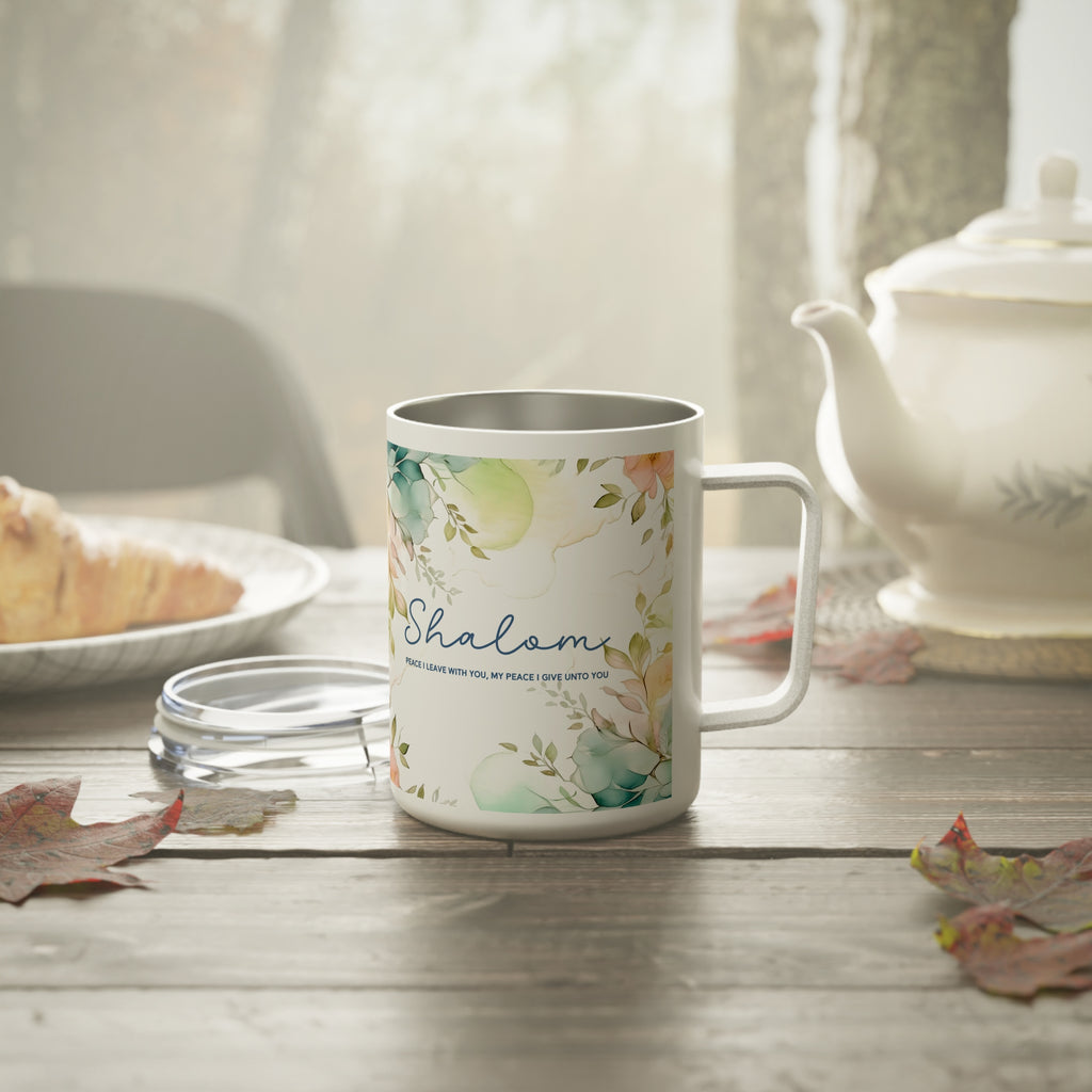 A white stainless steel -style mug with a floral watercolor print and the word 'Shalom' in blue script, set on a wooden table with a teapot, plate, and leaves in a softly lit kitchen or cafe scene.