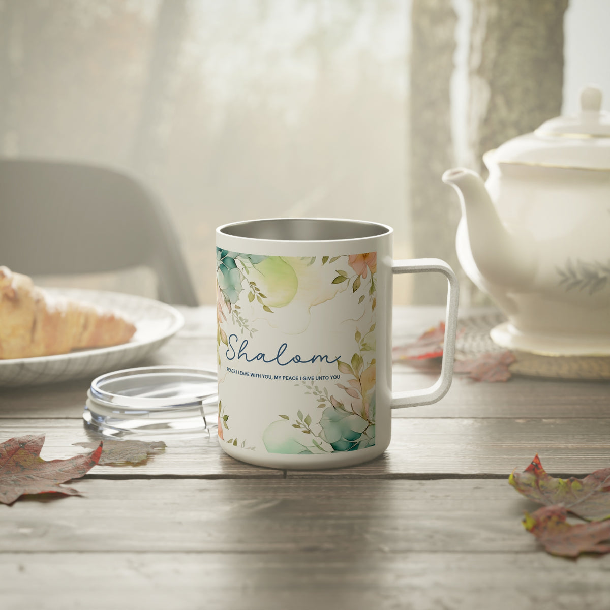 A white stainless steel -style mug with a floral watercolor print and the word 'Shalom' in blue script, set on a wooden table with a teapot, plate, and leaves in a softly lit kitchen or cafe scene.