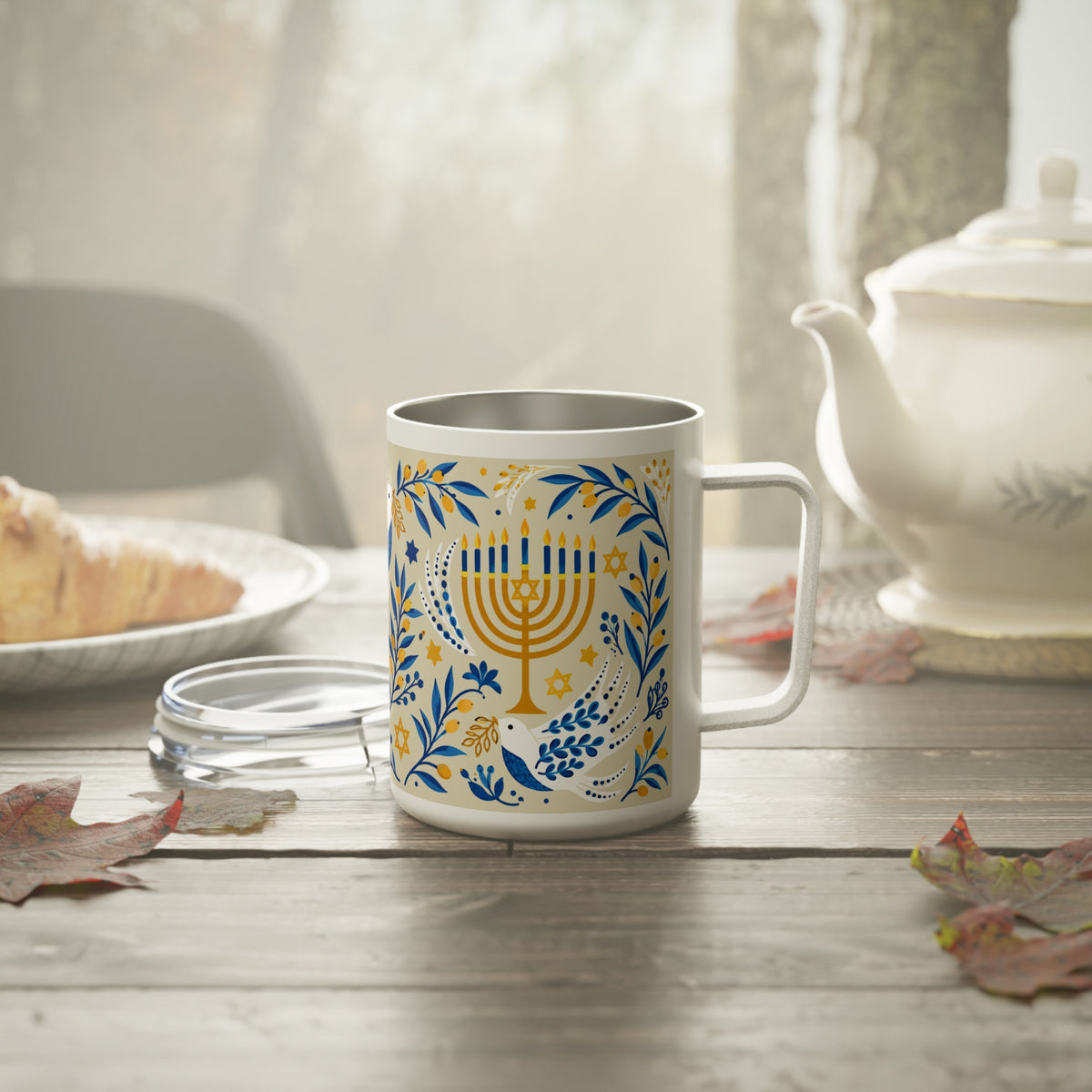 A white stainless steel mug with a gold and blue Hanukkah menorah and floral pattern, set on a rustic wooden table next to a glass lid, with a teapot and food blurred in the background.