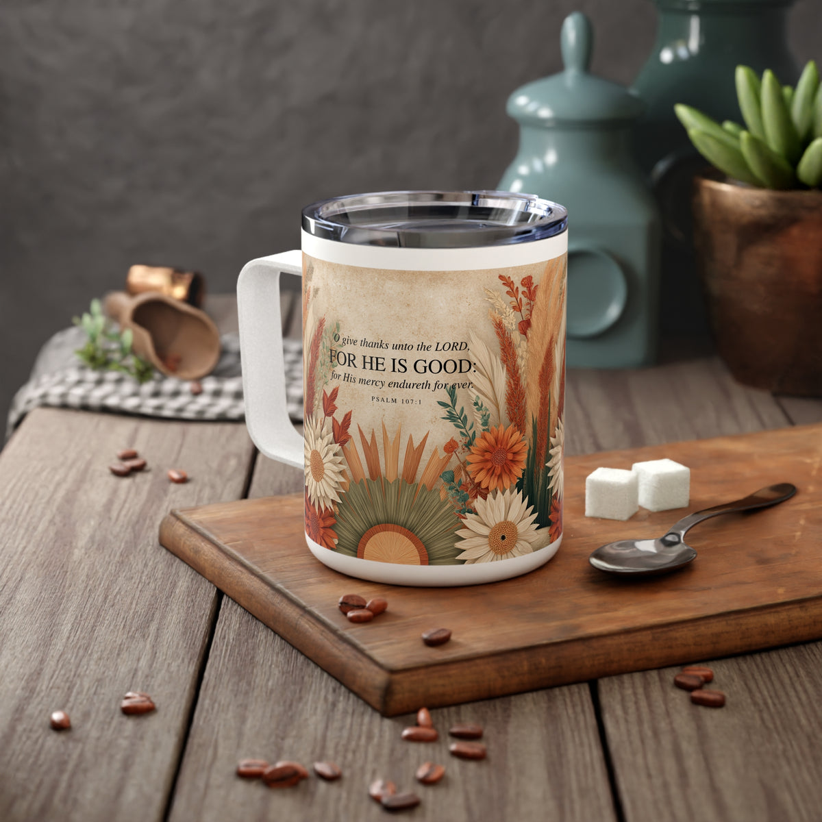 A white stainless steel mug with a colorful floral and botanical print featuring the Bible verse 'For He Is Good' (Psalm 34:8), styled on a wooden cutting board with coffee beans, a spoon, and sugar cubes in a rustic kitchen setting.