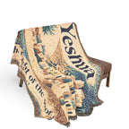 A woven throw blanket with fringe, draped over a brown bench against a plain white background. The blanket features a detailed tapestry-style design with the word 'Yeshua' in large dark blue letters, along with architectural imagery and additional text.