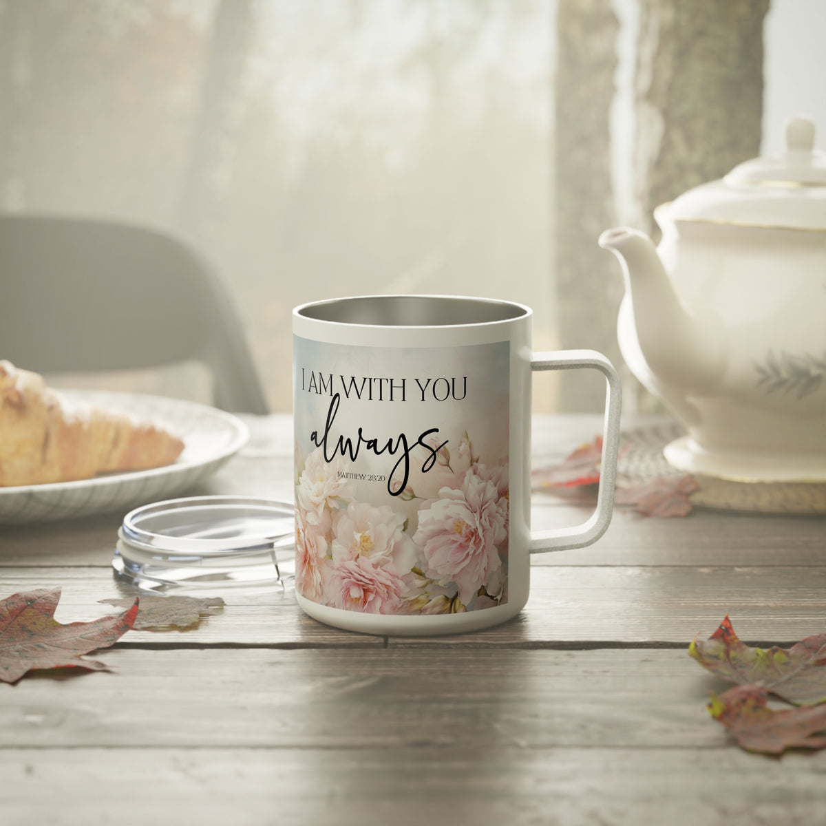 A white stainless steel mug with a floral design and the text 'I AM WITH YOU always' sits on a wooden table next to a teapot and plate, with a glass lid in front of it.
