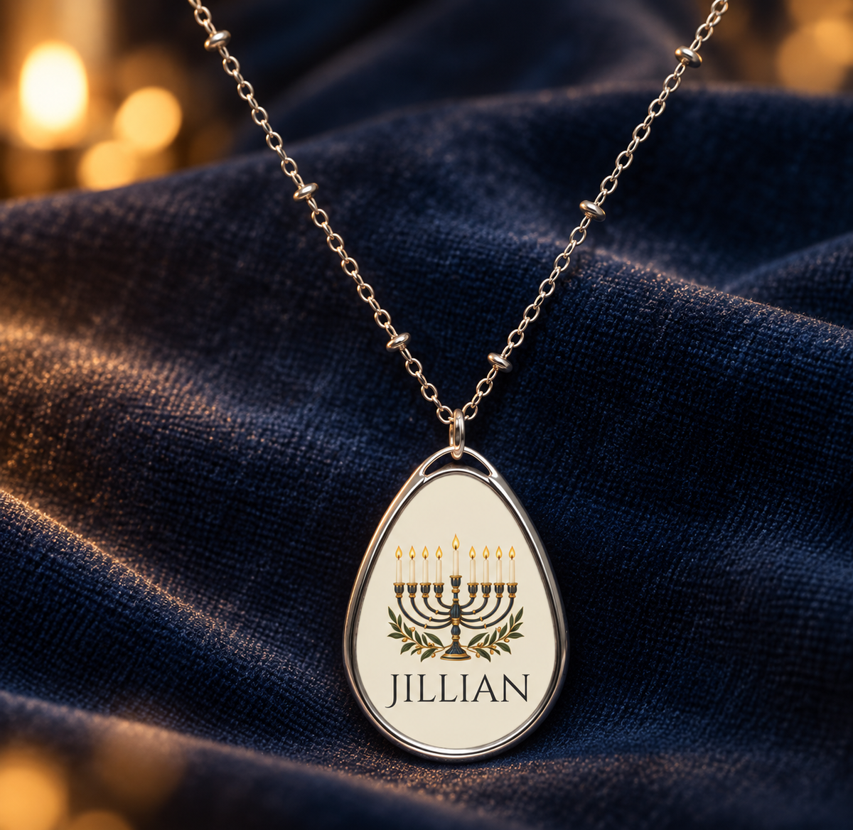 A silver teardrop pendant necklace with a menorah and the name 'JILLIAN' printed on it, resting on draped dark blue fabric with warm golden bokeh in the background.