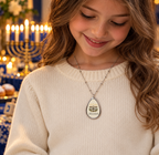 A young girl with long wavy brown hair smiles while wearing a silver teardrop pendant necklace with a menorah and the name 'HILARY' engraved, set against a warm, festive background with candles and a blue-and-white table setting.