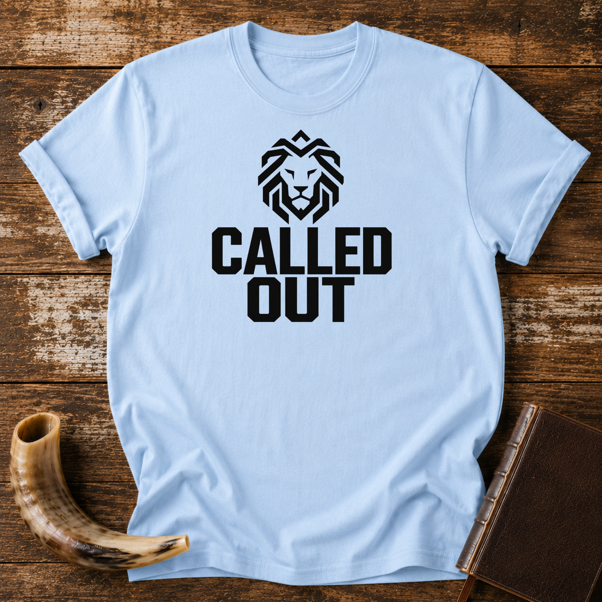 Light blue t-shirt with a geometric black lion head over bold CALLED OUT text, laid flat with a shofar and a leather book