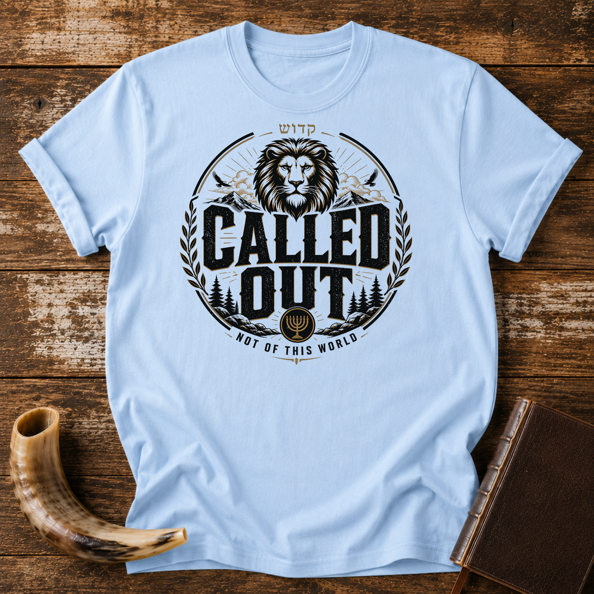 Light blue t-shirt with a CALLED OUT lion badge with Hebrew lettering, a menorah and NOT OF THIS WORLD, laid flat with a shofar and a leather book