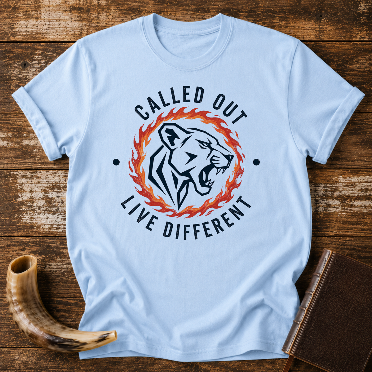 Light blue t-shirt with a roaring lioness in a ring of fire with CALLED OUT and LIVE DIFFERENT, laid flat with a shofar and a leather book