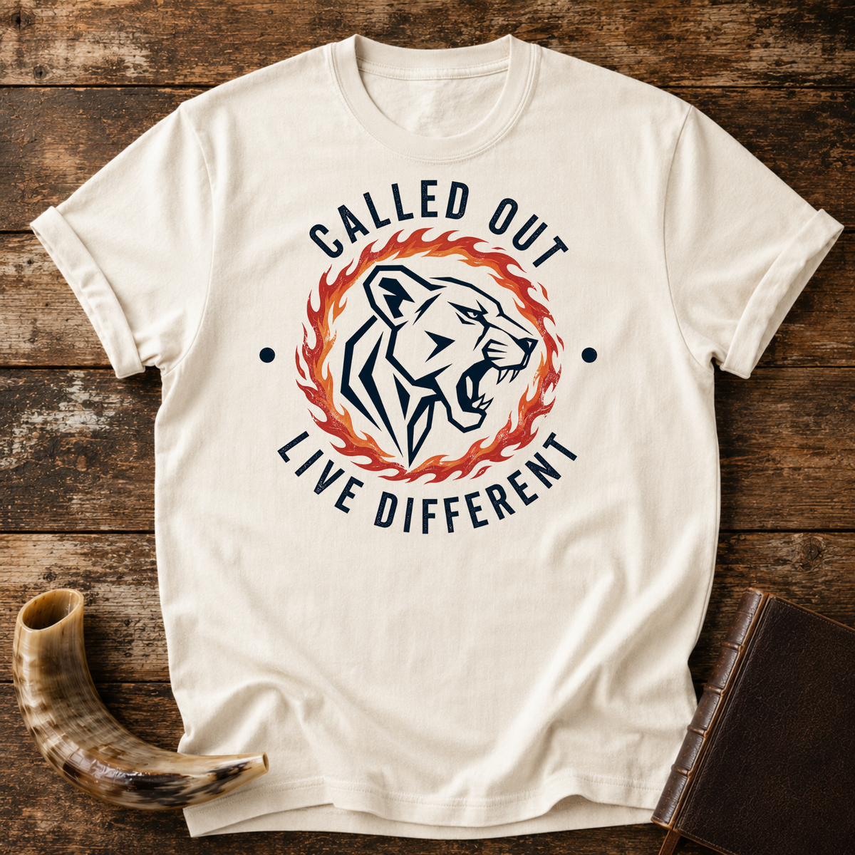 Natural cream t-shirt with a roaring lioness in a ring of fire with CALLED OUT and LIVE DIFFERENT, laid flat with a shofar and a leather book