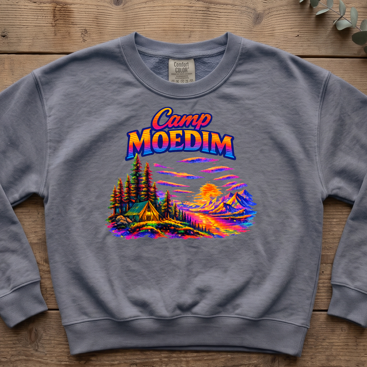 Blue jean Camp Moedim sweatshirt with a bright neon tent, pine trees and sunset mountain scene, laid flat on wood boards