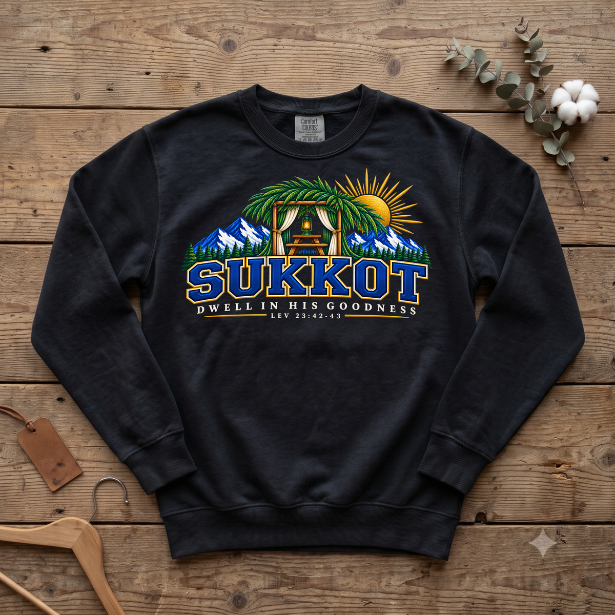 Black Sukkot Dwell in His Goodness Lev 23:42-43 sweatshirt with a sukkah, mountains and sun, laid flat on wood with a hanger and cotton stem