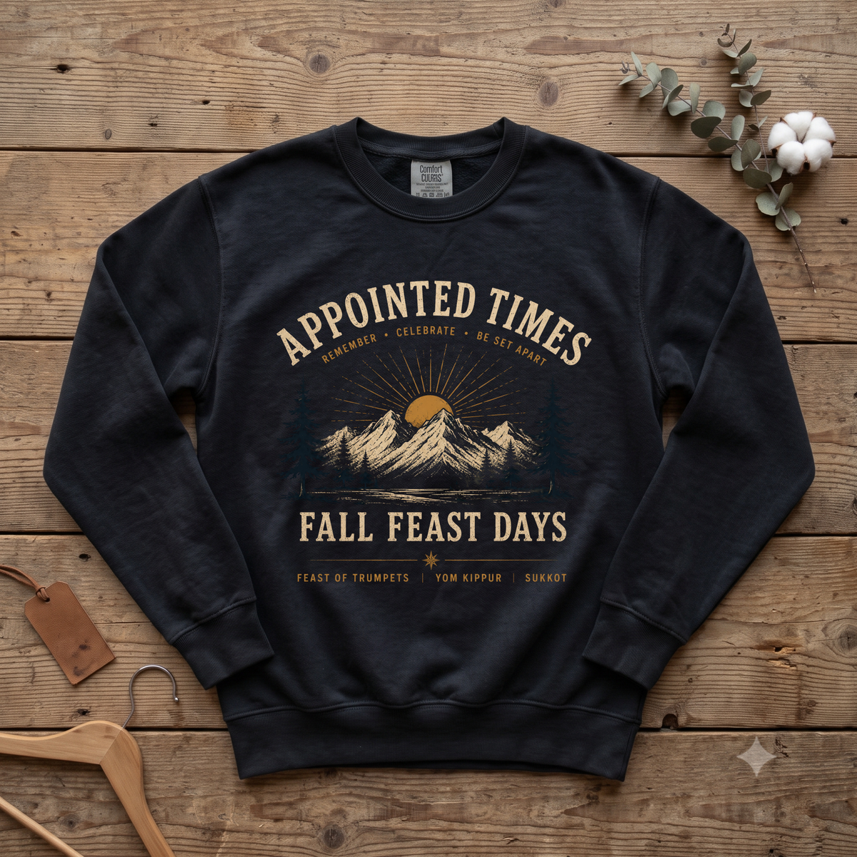 Black Appointed Times Fall Feast Days sweatshirt with a mountain sunrise and pines, laid flat on wood with a hanger and cotton stem