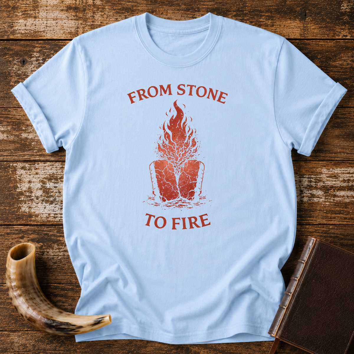 Light blue From Stone to Fire tee with red flames rising from stone tablets, laid flat with a shofar and a leather-bound book