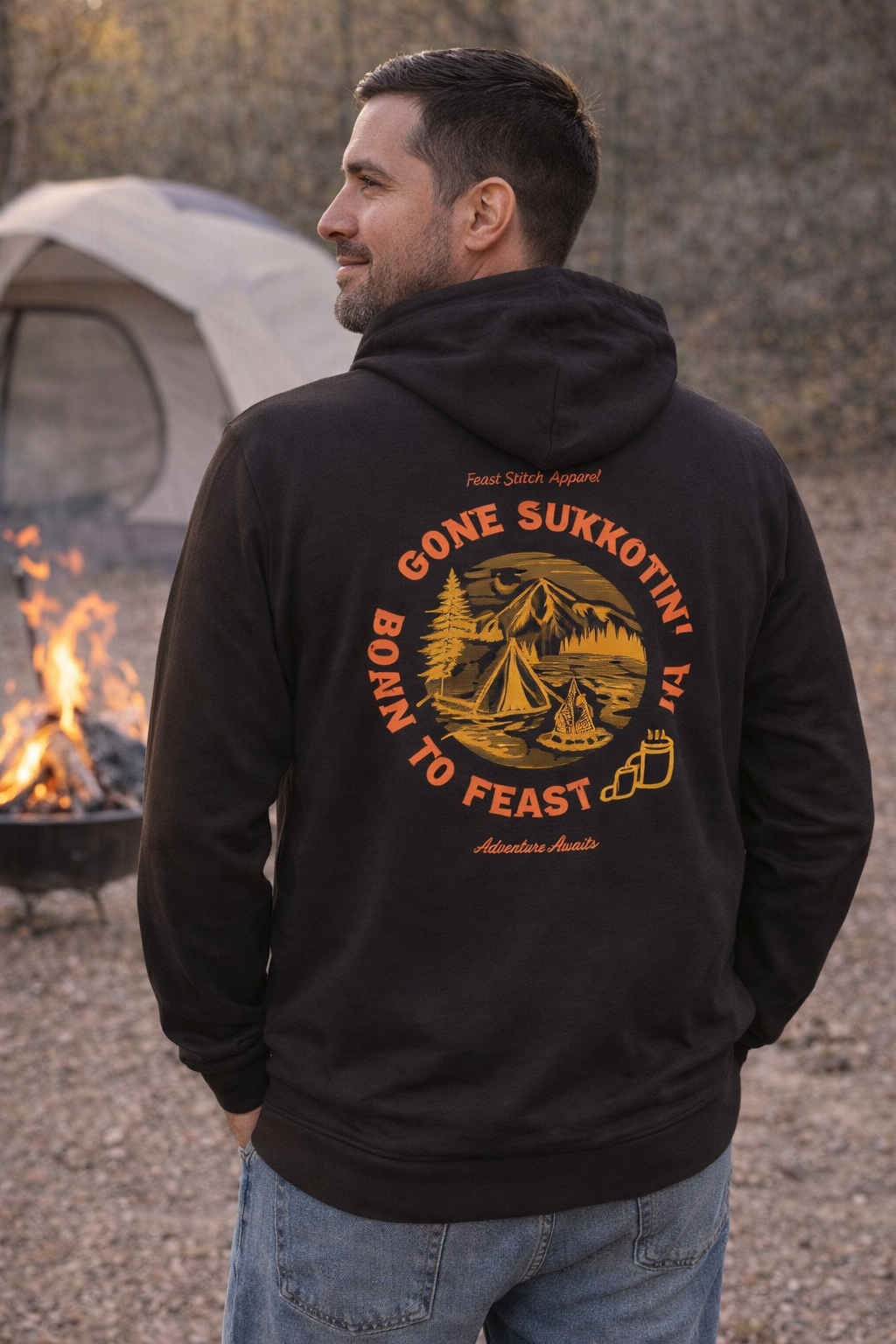 Back of a black Gone Sukkotin' Born to Feast hoodie with an orange camping design, worn by a man at a campsite