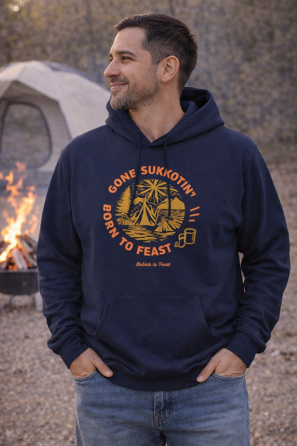Navy Gone Sukkotin' Born to Feast hoodie with an orange and gold camping design, worn by a smiling man by a campfire