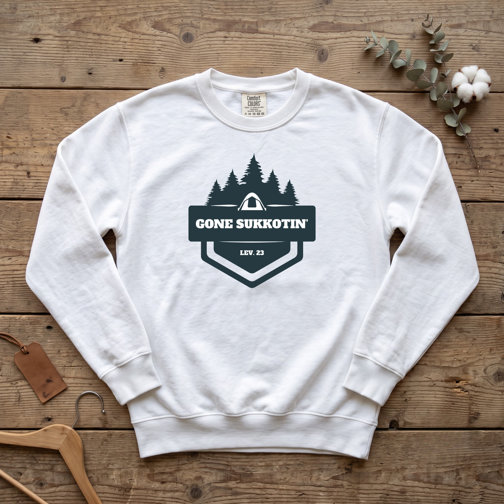 White Gone Sukkotin' Lev. 23 sweatshirt with a badge of pine trees and a tent, laid flat on a wooden table