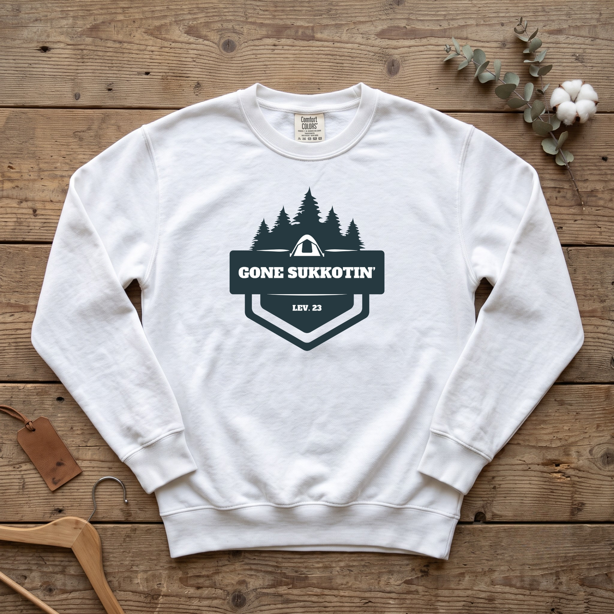 White Gone Sukkotin' Lev. 23 sweatshirt with a badge of pine trees and a tent, laid flat on a wooden table