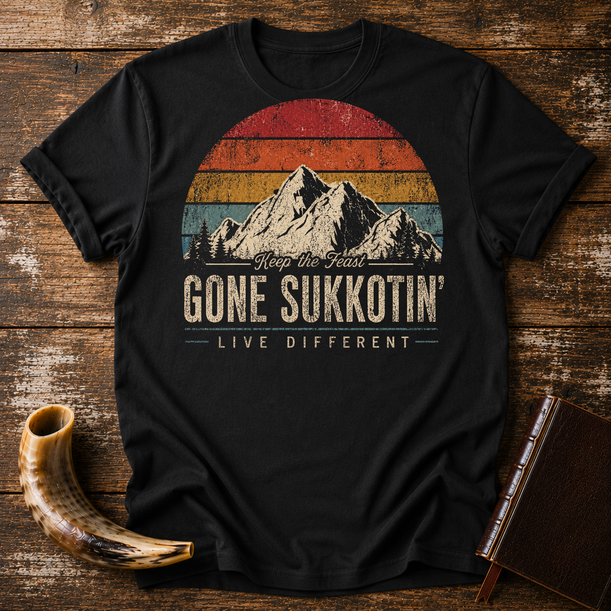 Black t-shirt with GONE SUKKOTIN' LIVE DIFFERENT under mountains and a retro sunset, laid flat with a shofar and a leather book