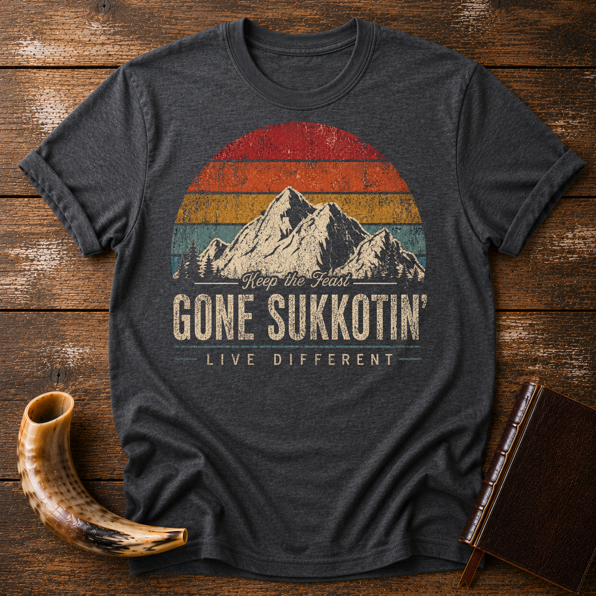 Dark heather gray t-shirt with GONE SUKKOTIN' LIVE DIFFERENT under mountains and a retro sunset, laid flat with a shofar and a leather book