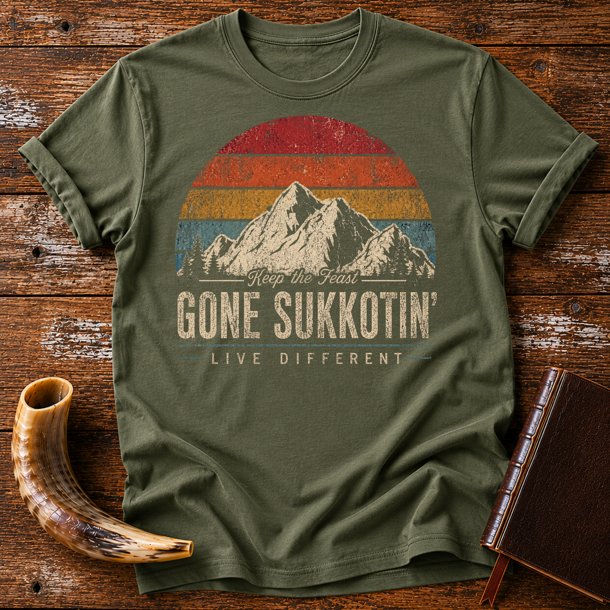 Military green t-shirt with GONE SUKKOTIN' LIVE DIFFERENT under mountains and a retro sunset, laid flat with a shofar and a leather book
