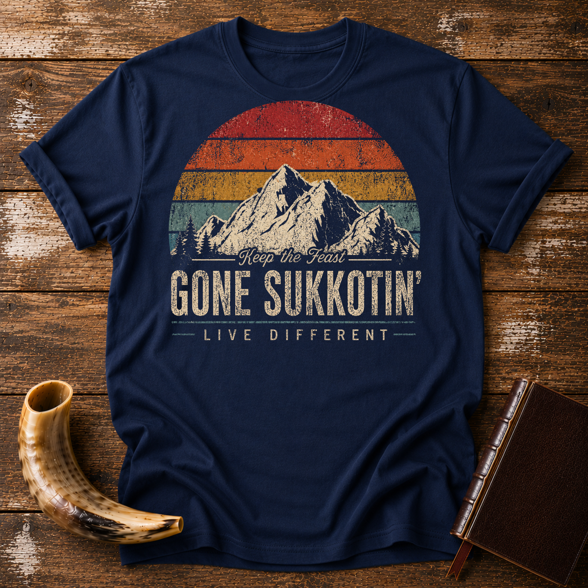 Navy t-shirt with GONE SUKKOTIN' LIVE DIFFERENT under mountains and a retro sunset, laid flat with a shofar and a leather book