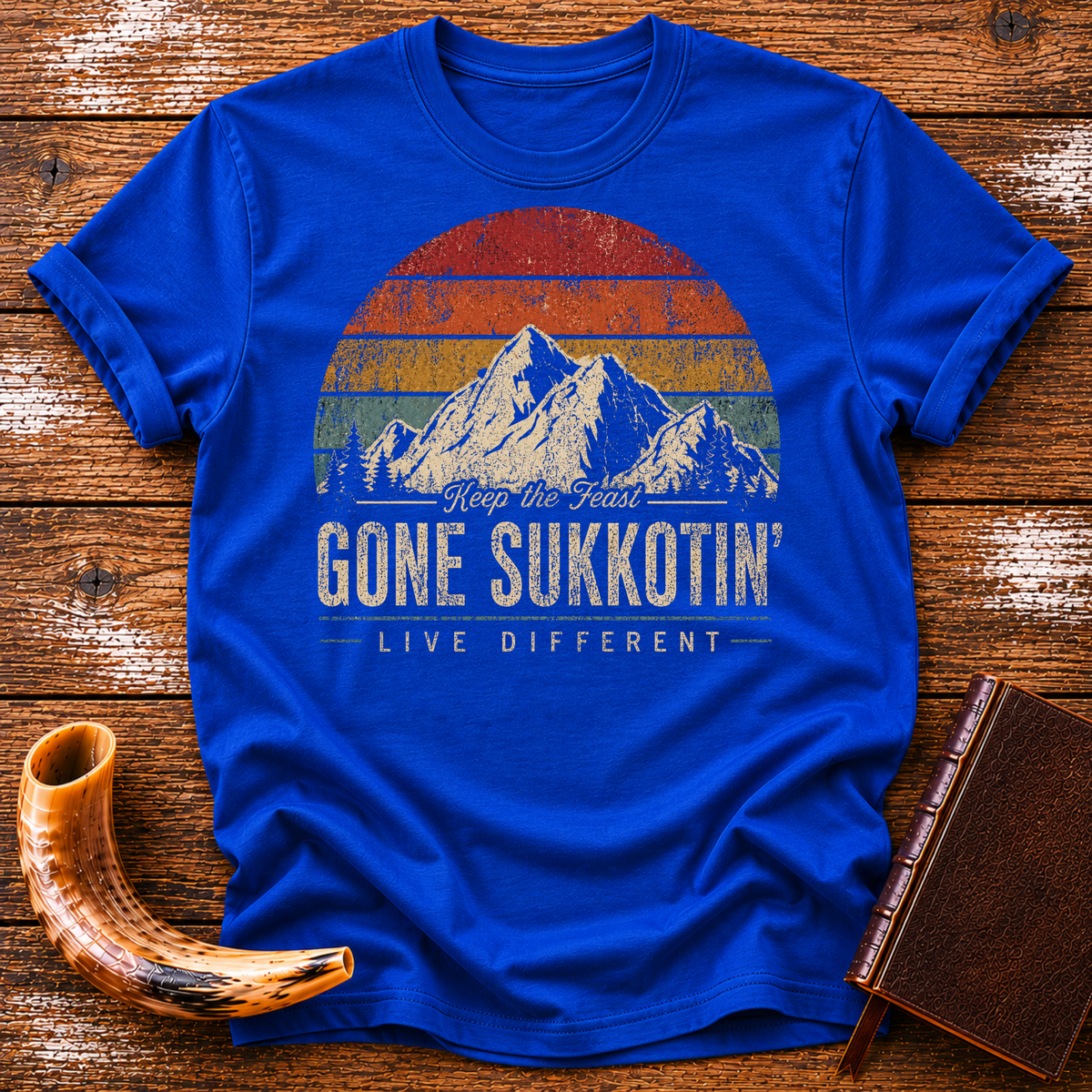 Royal blue t-shirt with GONE SUKKOTIN' LIVE DIFFERENT under mountains and a retro sunset, laid flat with a shofar and a leather book