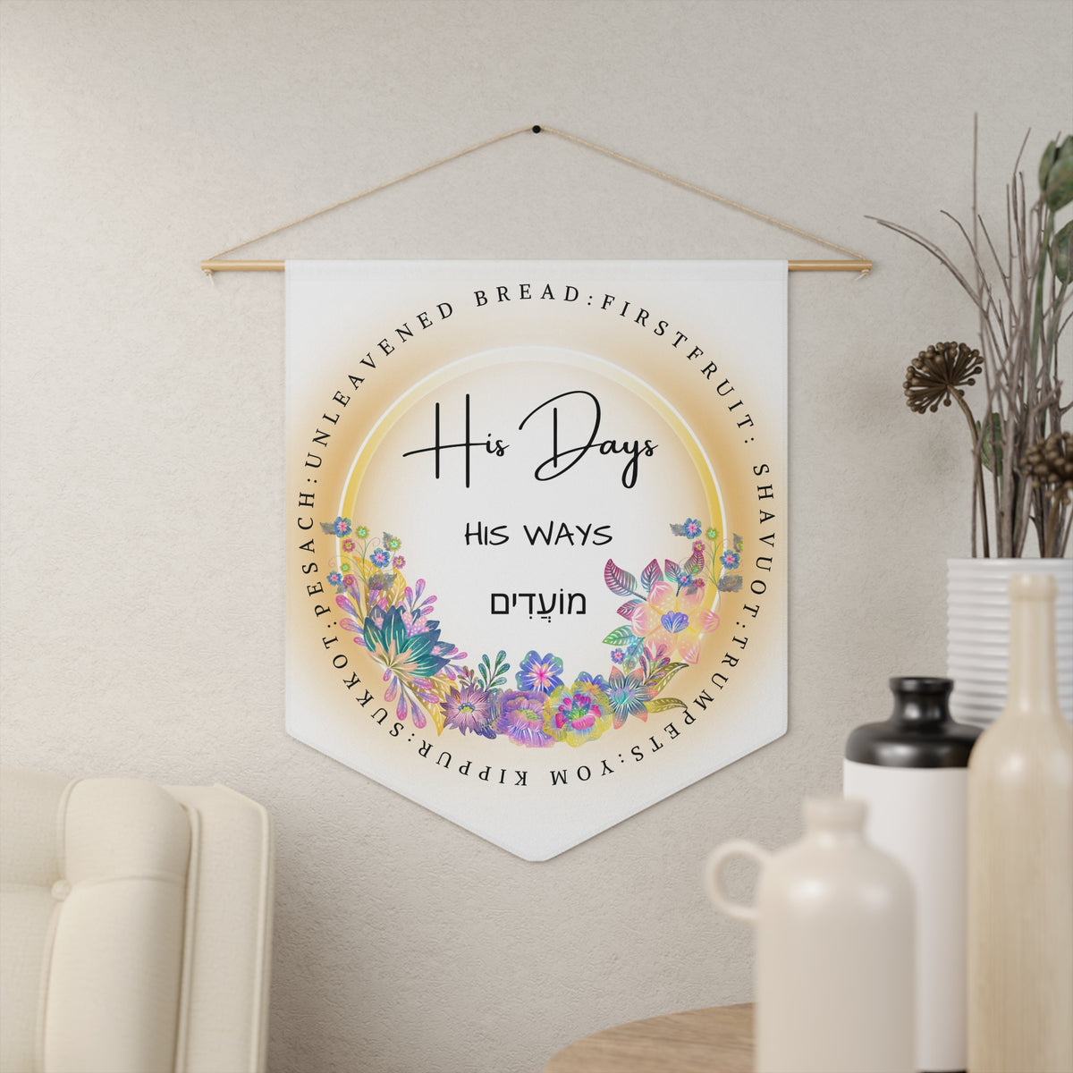 His Days His Ways wall pennant with Hebrew lettering in a floral wreath ringed by feast day names, hanging on a cream wall beside vases