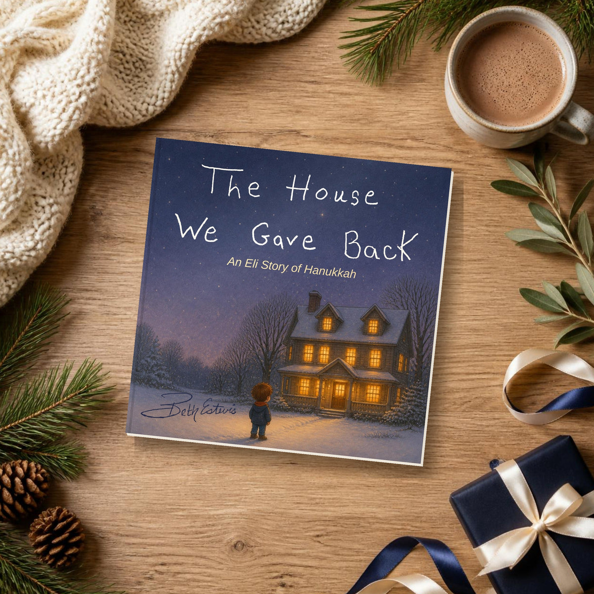 The House We Gave Back Hanukkah picture book on a wooden table with a knit blanket, hot cocoa, pine sprigs and a navy gift box