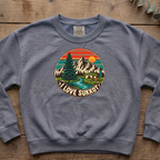 Blue jean I Love Sukkot sweatshirt with a round mountain, pine tree and river camping scene, laid flat on a wooden table