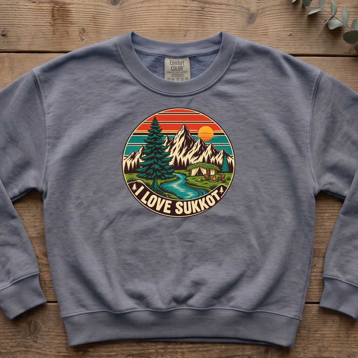 Blue jean I Love Sukkot sweatshirt with a round mountain, pine tree and river camping scene, laid flat on a wooden table