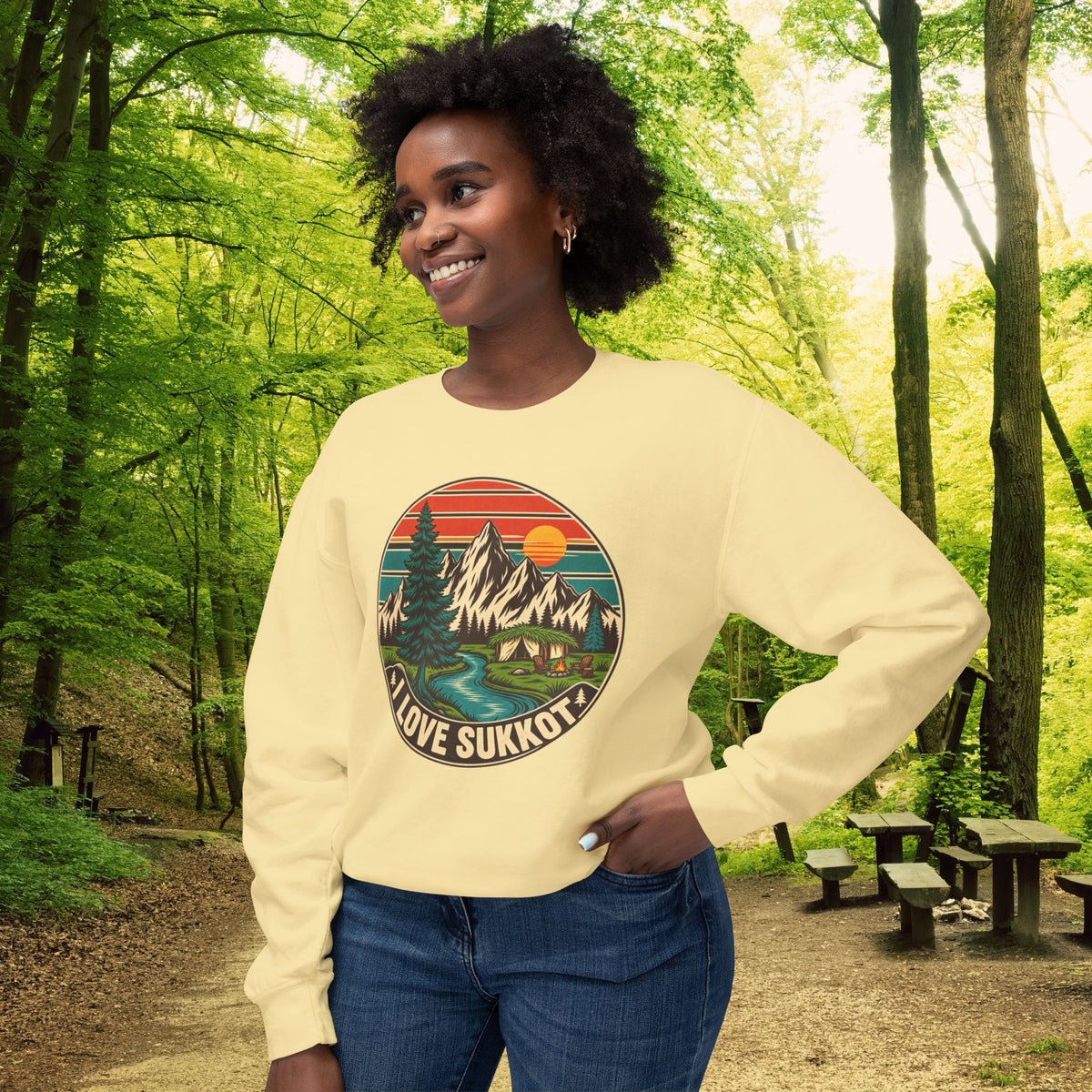 Butter I Love Sukkot sweatshirt with a round mountain, pine tree and river camping scene, worn by a smiling woman in a forest