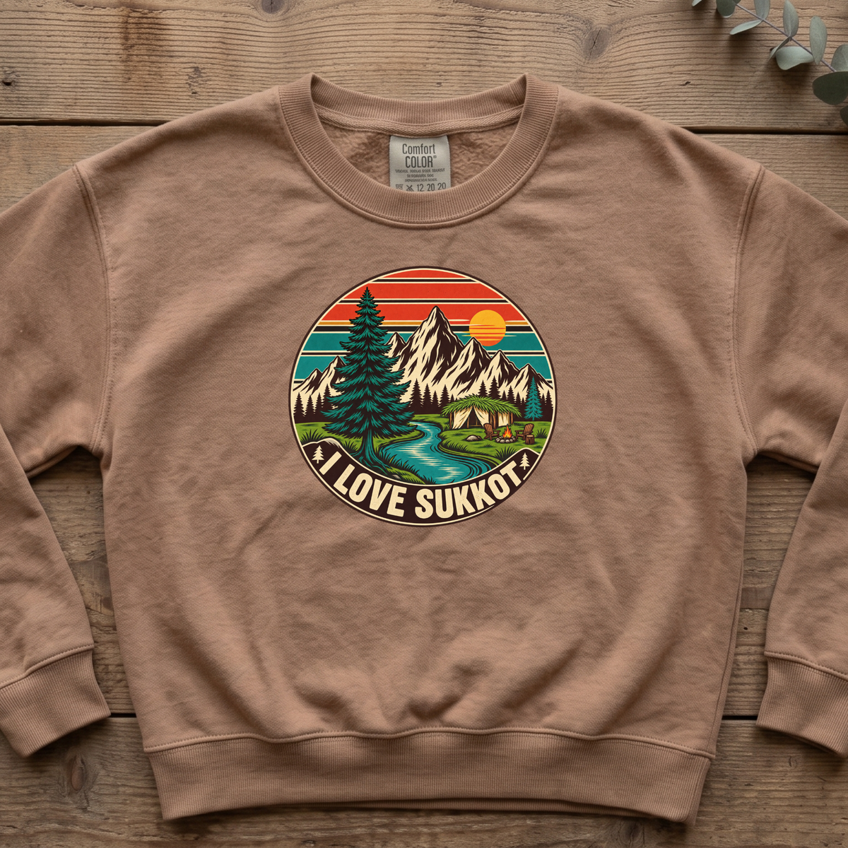 Espresso I Love Sukkot sweatshirt with a round mountain, pine tree and river camping scene, laid flat on a wooden table