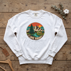 White I Love Sukkot sweatshirt with a round mountain, pine tree and river camping scene, laid flat on a wooden table