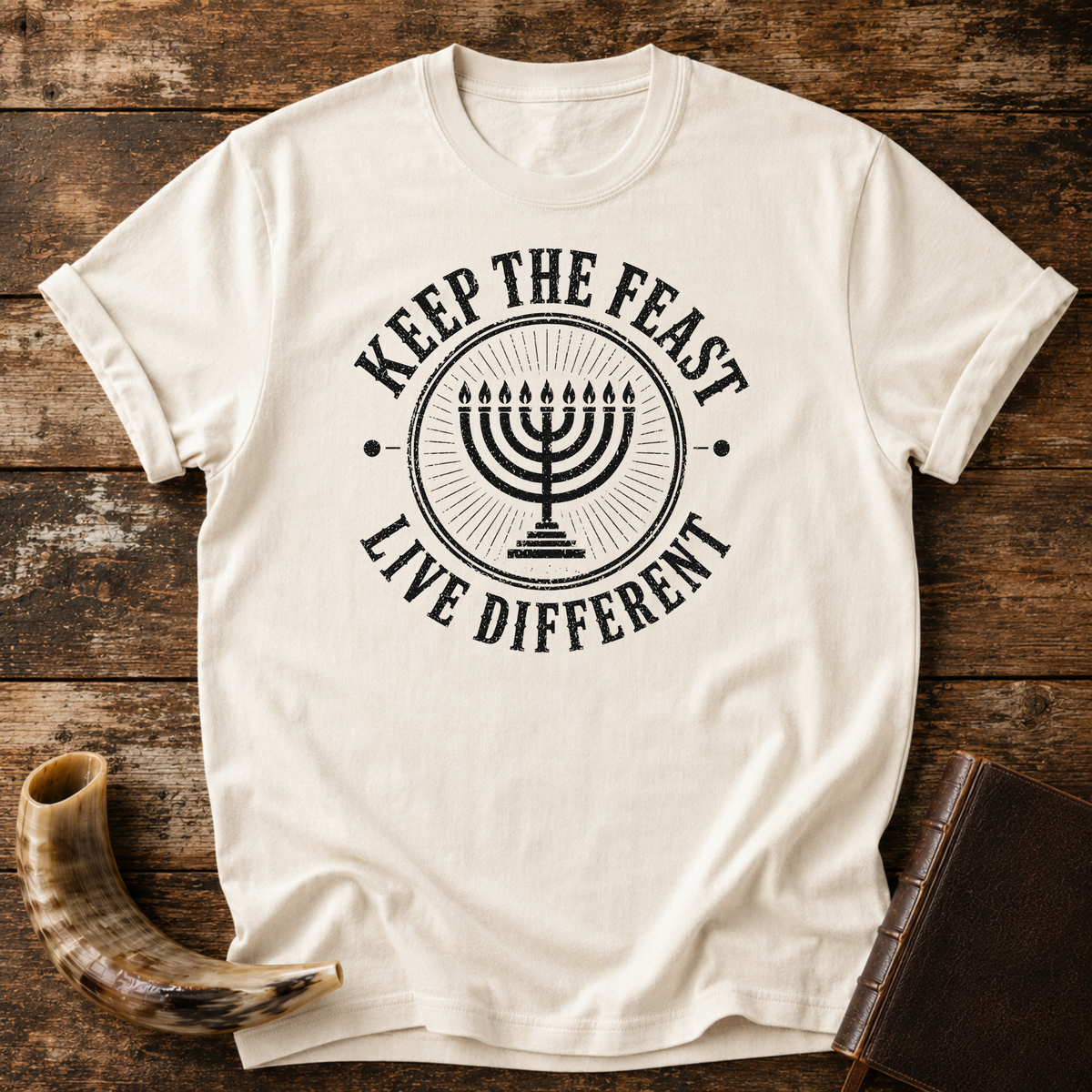 Natural cream t-shirt with a black seven-branch menorah inside KEEP THE FEAST LIVE DIFFERENT, laid flat with a shofar and a leather book