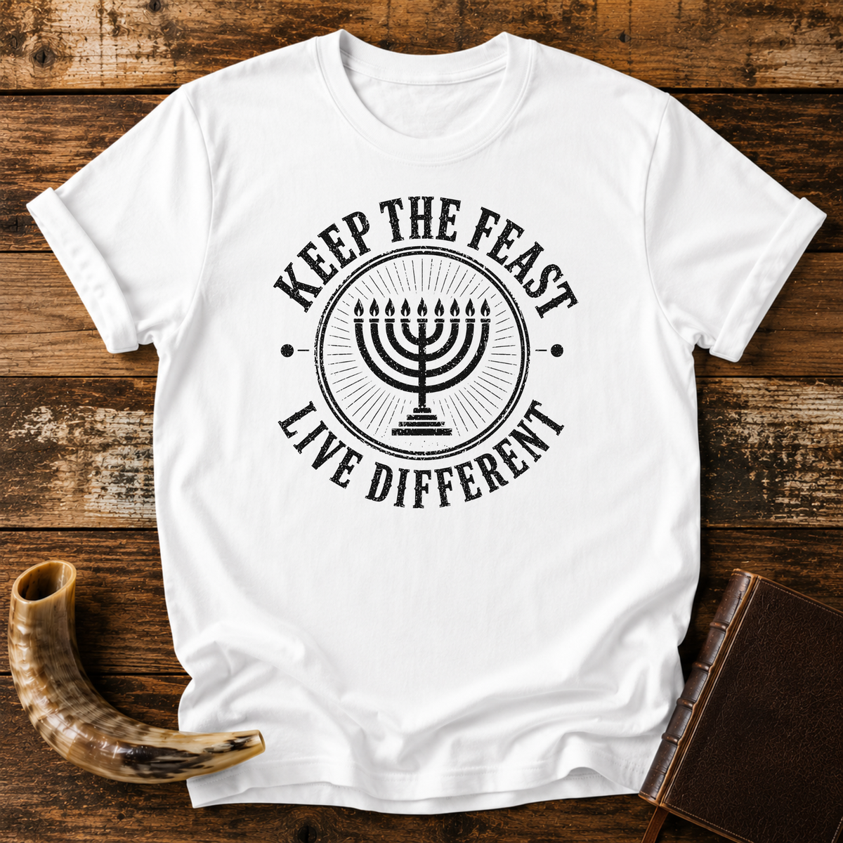 White t-shirt with a black seven-branch menorah inside KEEP THE FEAST LIVE DIFFERENT, laid flat with a shofar and a leather book