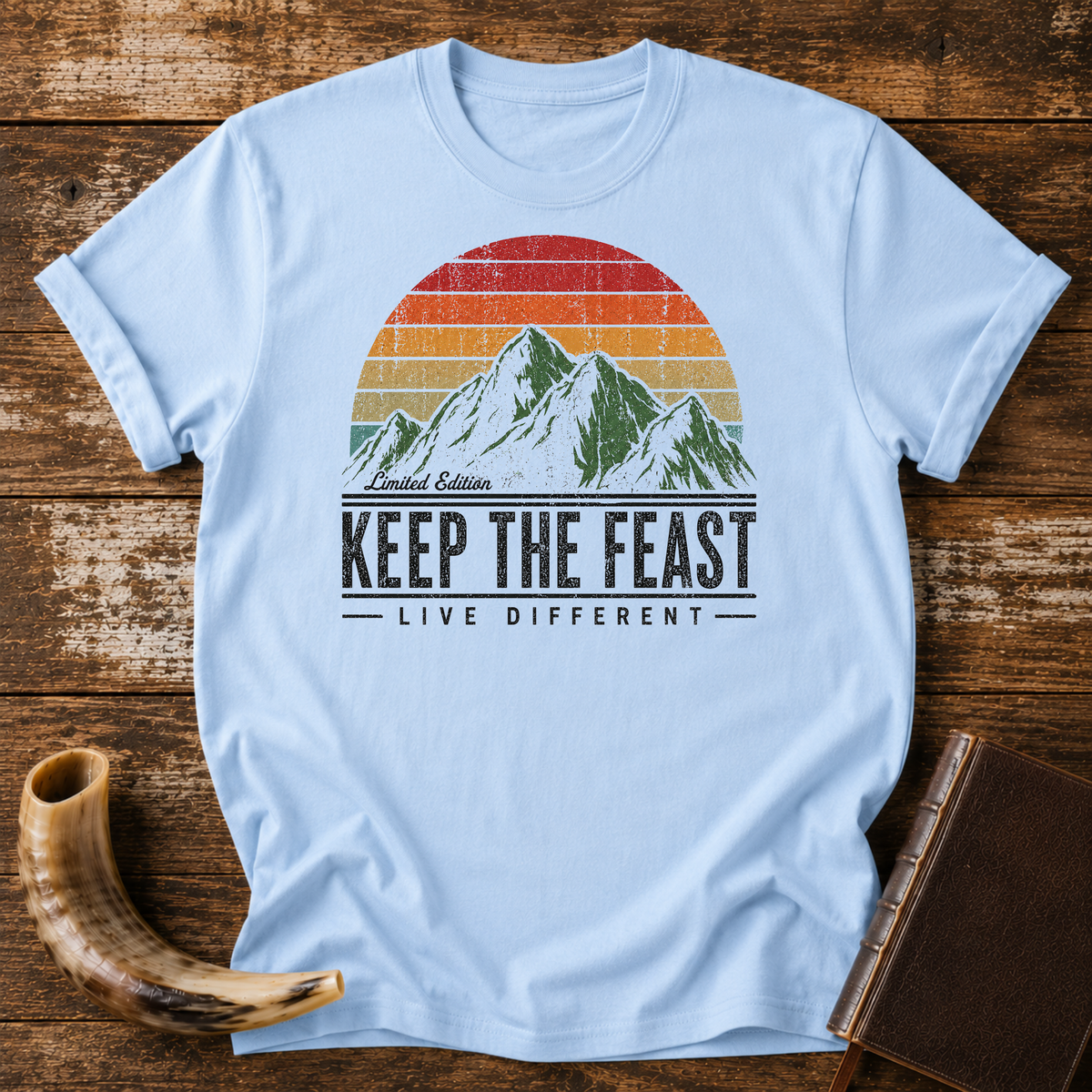 Light blue t-shirt with Limited Edition KEEP THE FEAST LIVE DIFFERENT and a retro mountain sunset, laid flat with a shofar and a leather book