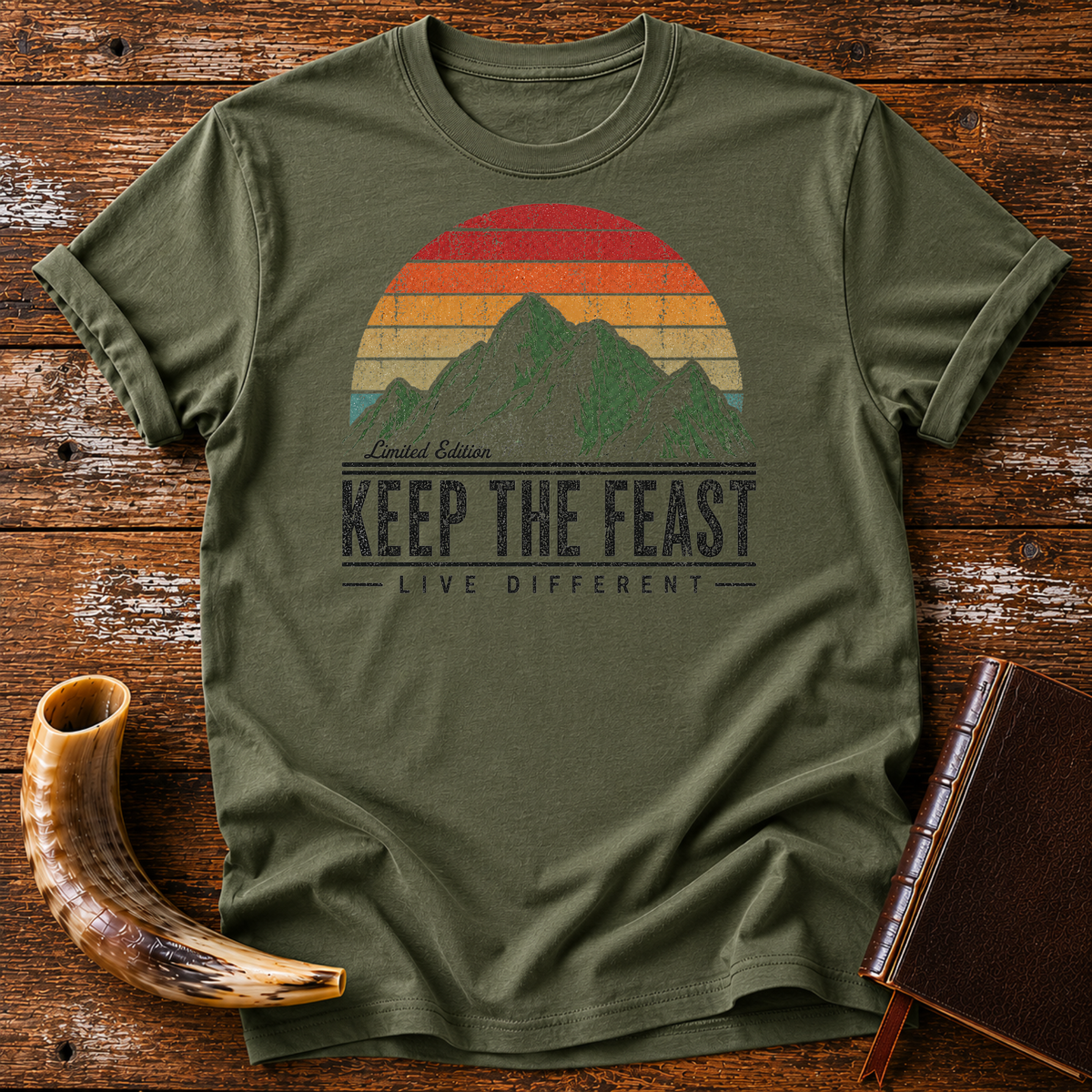 Military green t-shirt with Limited Edition KEEP THE FEAST LIVE DIFFERENT and a retro mountain sunset, laid flat with a shofar and a leather book