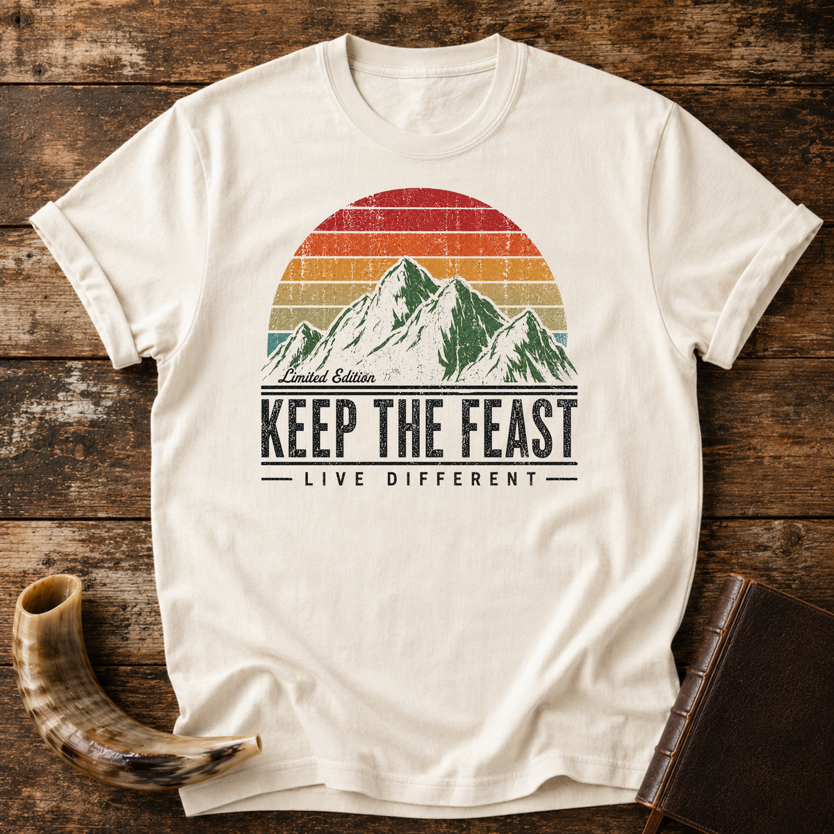 Natural cream t-shirt with Limited Edition KEEP THE FEAST LIVE DIFFERENT and a retro mountain sunset, laid flat with a shofar and a leather book