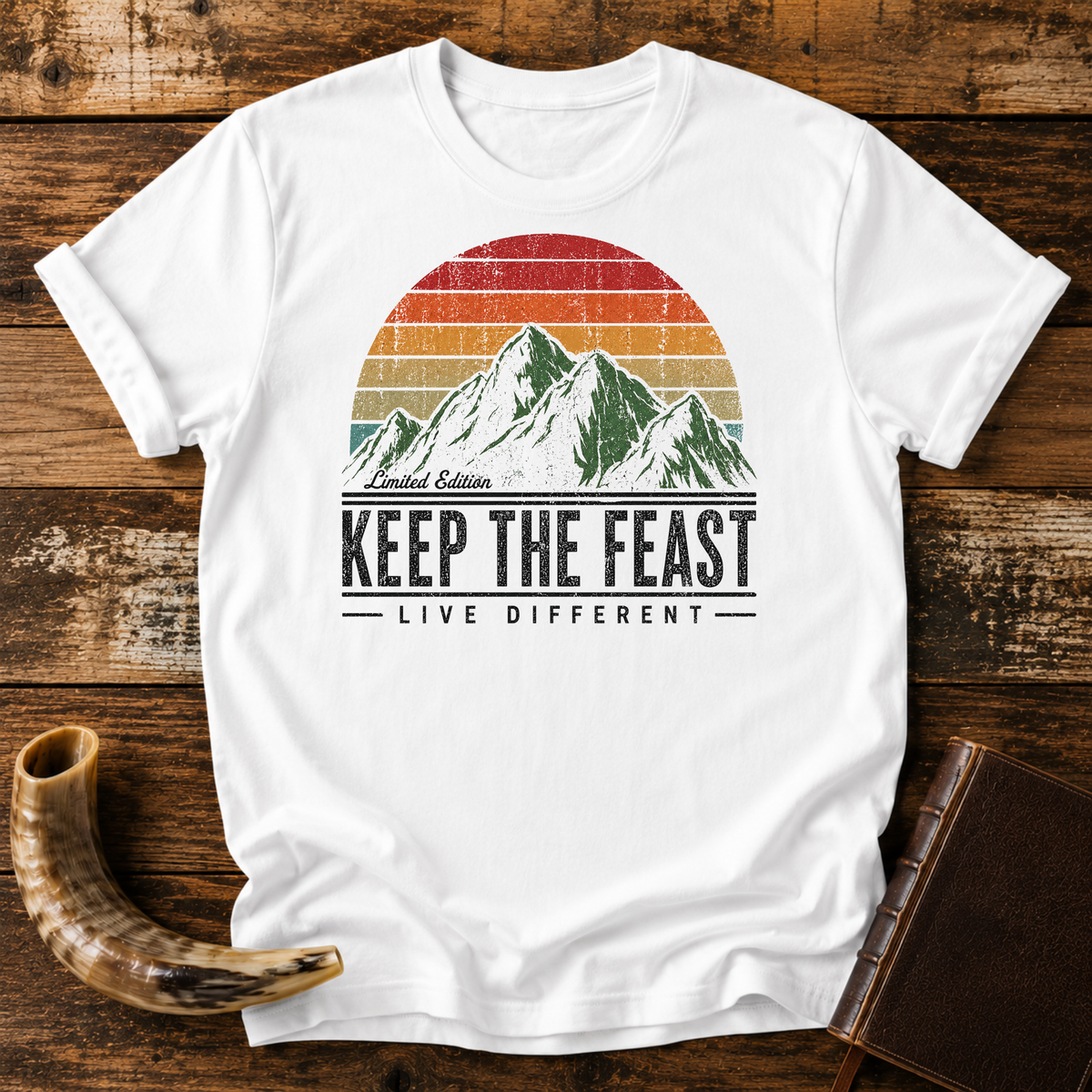 White t-shirt with Limited Edition KEEP THE FEAST LIVE DIFFERENT and a retro mountain sunset, laid flat with a shofar and a leather book