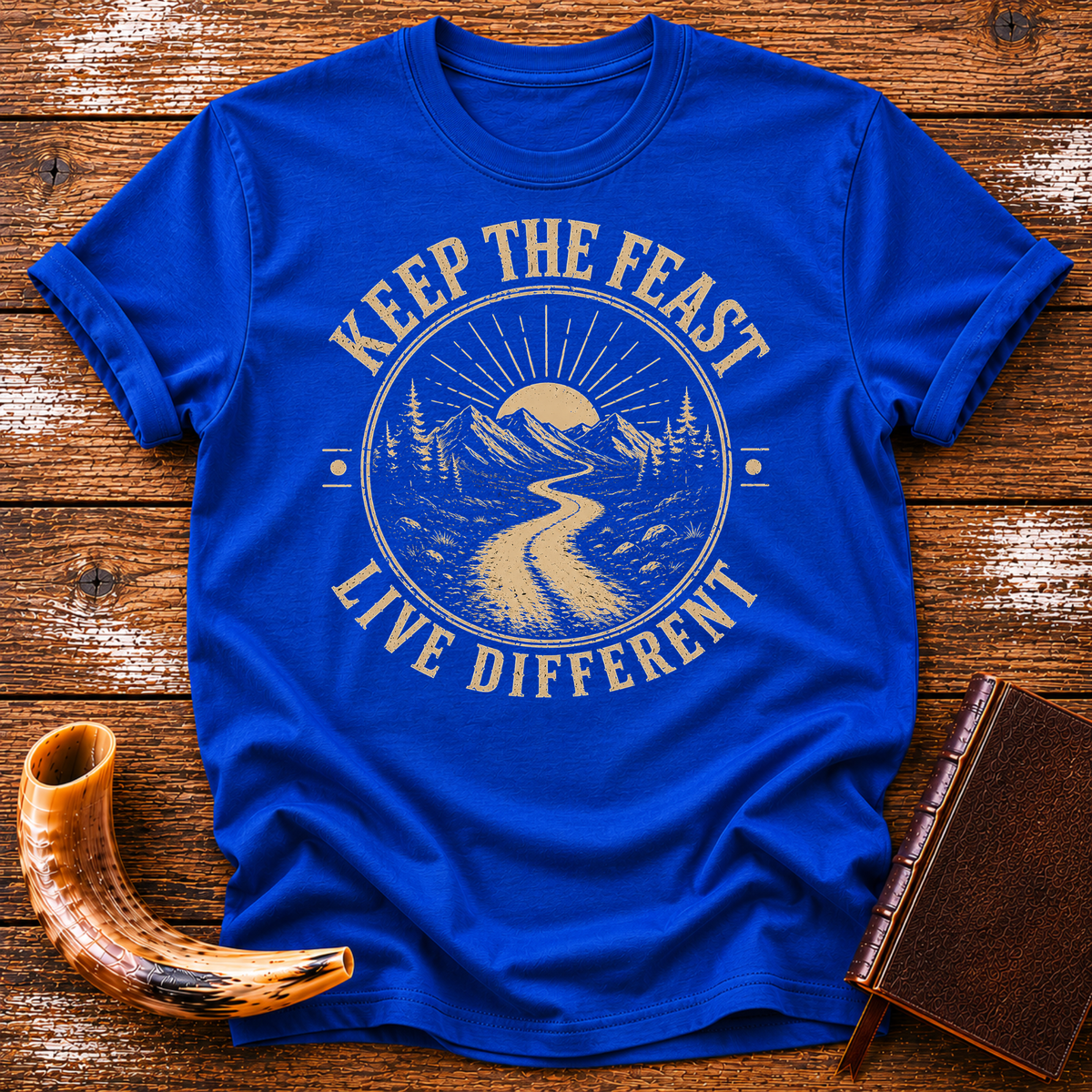 Royal blue t-shirt with KEEP THE FEAST LIVE DIFFERENT around a winding road toward a rising sun, laid flat with a shofar and a leather book
