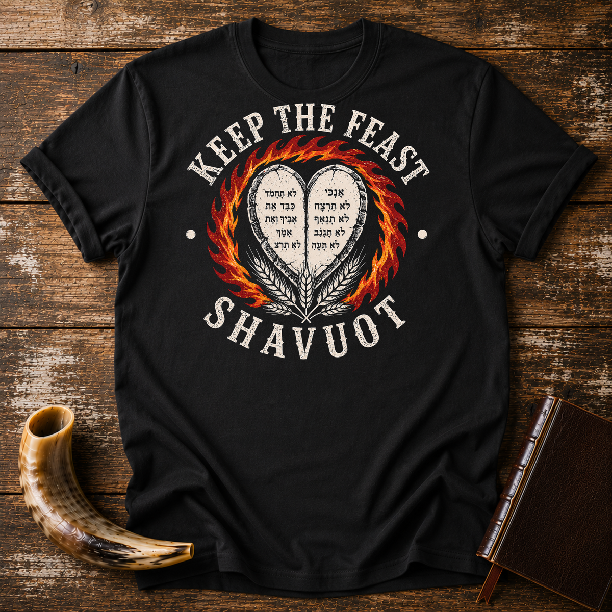Black t-shirt with KEEP THE FEAST SHAVUOT in cream, heart tablets, fire and wheat, laid flat with a shofar and a leather book