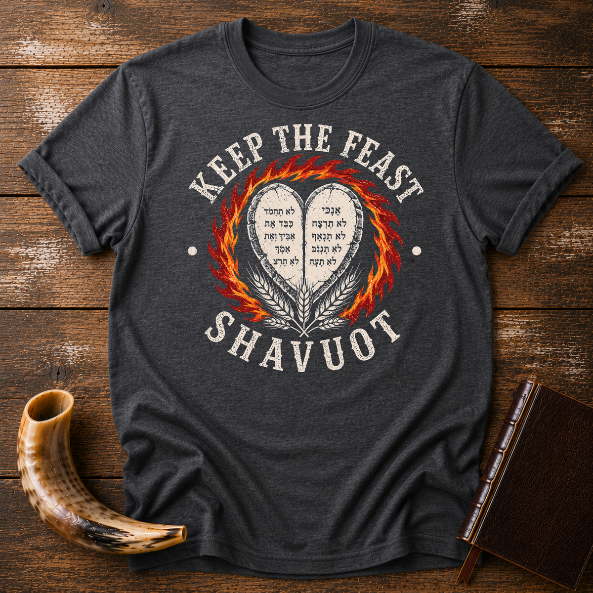 Dark heather gray t-shirt with KEEP THE FEAST SHAVUOT in cream, heart tablets, fire and wheat, laid flat with a shofar and a leather book