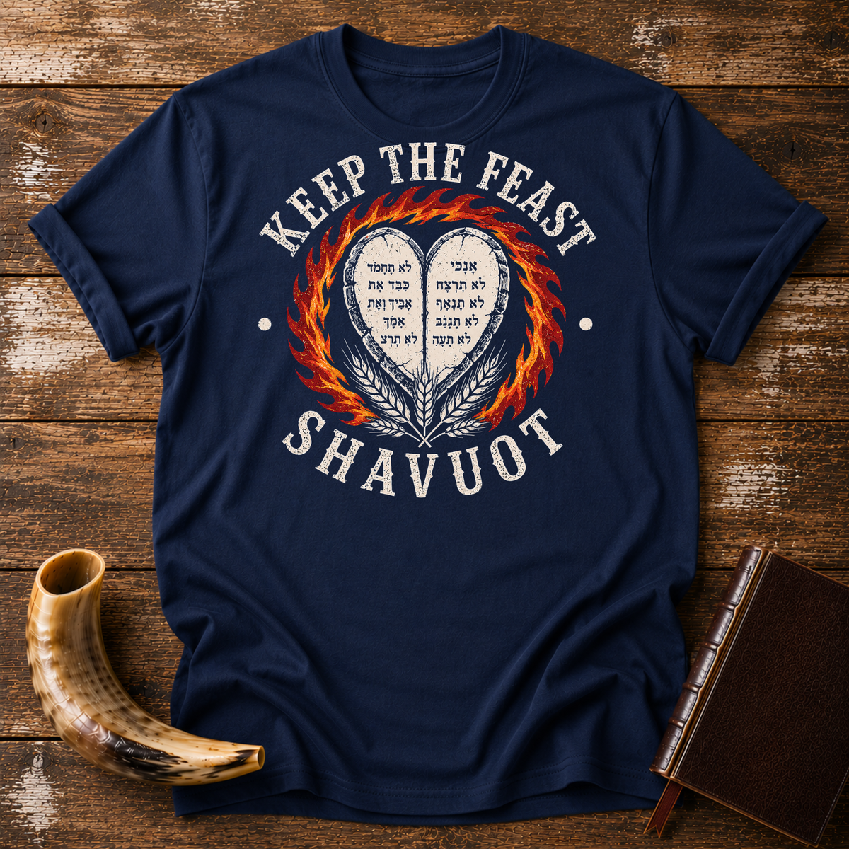 Navy t-shirt with KEEP THE FEAST SHAVUOT in cream, heart tablets, fire and wheat, laid flat with a shofar and a leather book