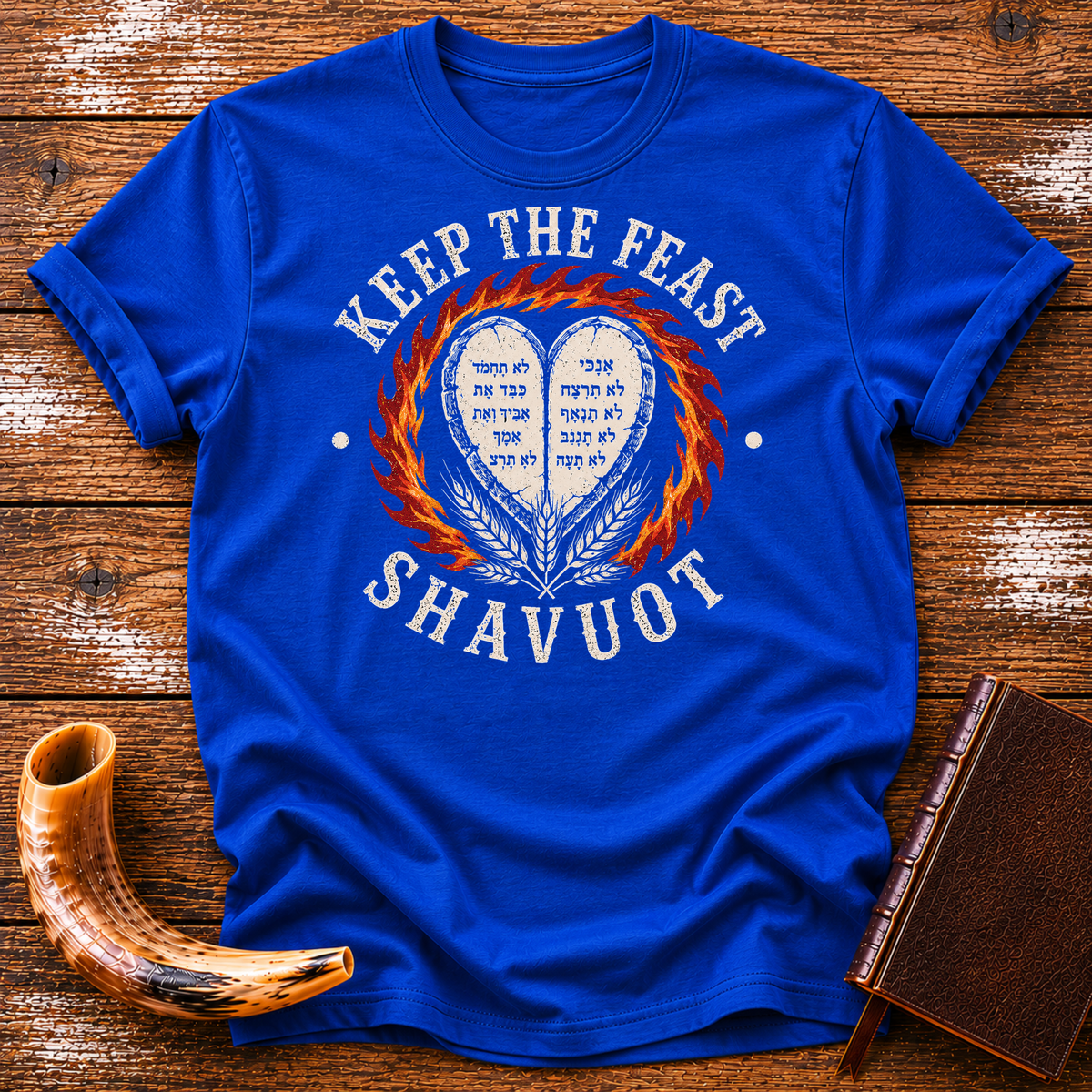 Royal blue t-shirt with KEEP THE FEAST SHAVUOT in cream, heart tablets, fire and wheat, laid flat with a shofar and a leather book