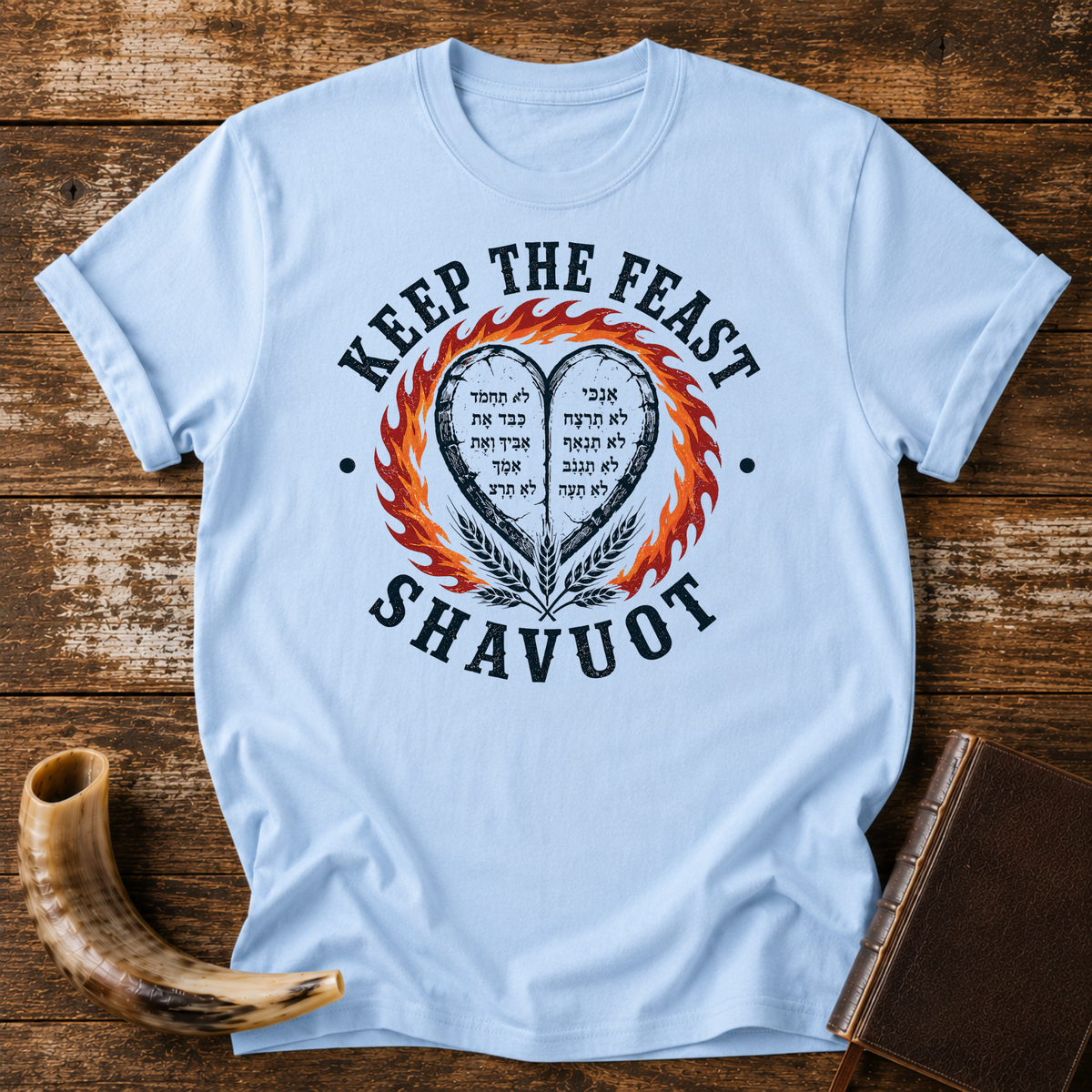Light blue t-shirt with KEEP THE FEAST SHAVUOT, Hebrew-lettered heart tablets, fire and wheat, laid flat with a shofar and a leather book