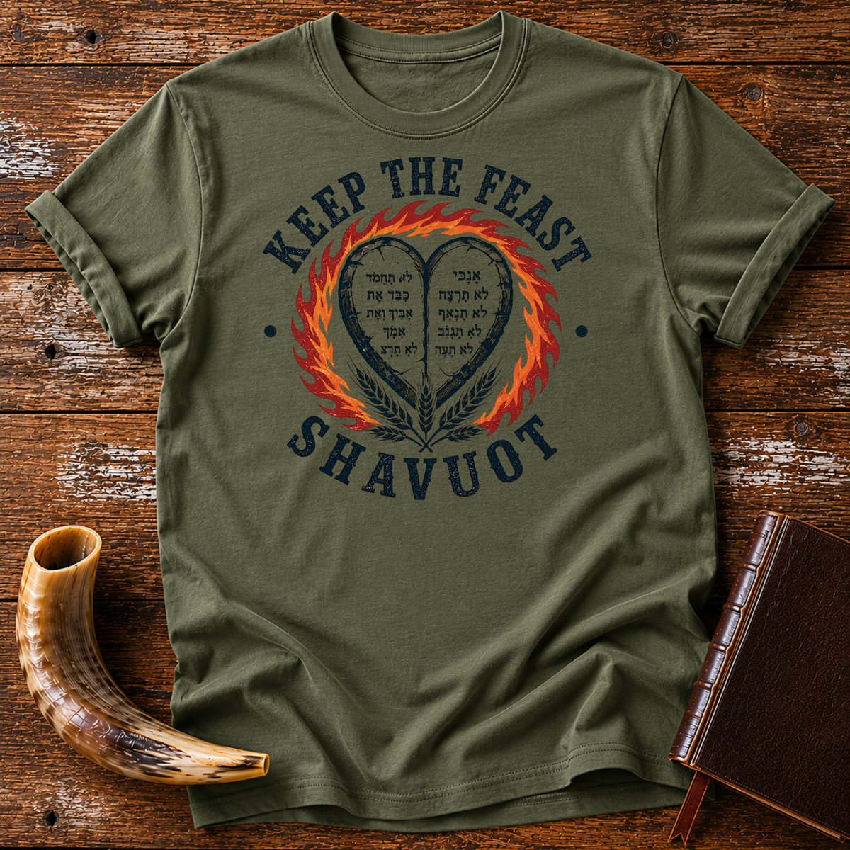 Military green t-shirt with KEEP THE FEAST SHAVUOT, Hebrew-lettered heart tablets, fire and wheat, laid flat with a shofar and a leather book